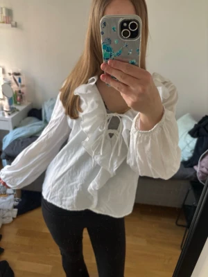Vit volangblus med knytning - Supersöt vit blus med volangdetaljer och knytning framtill. Blusen har långa, puffiga ärmar med resår vid handlederna och en djupare urringning. Perfekt för dig som gillar romantisk och trendig stil. Materialet känns lätt och luftigt. Både fin och skön blus, aldrig använd ändast testat på. Lapp kvar. 
