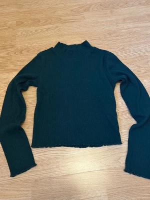 Mörkgrön ribbstickad långärmad tröja - Grön topp med lång ärm. Tröjan har vågiga kanter vid ärmslut och nederkant