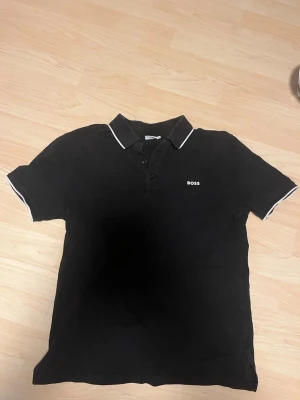 Svart pikétröja från Hugo BOSS - Svart pikétröja från BOSS med vit kant på krage och ärmslut. Klassisk design med knappar framtill och broderad BOSS-logga på bröstet. Perfekt för en stilren look och enkel att matcha med jeans eller shorts. Storlek 16 år barn .