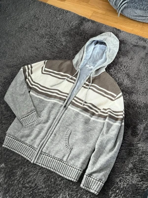  stickad cardigan  - Stickad hoodie i grått med dragkedja, huva och fickor framtill. Tröjan har breda bruna och vita ränder över bröstet och ryggen samt ribbade muddar och nederkant. Perfekt för dig som gillar en avslappnad och stilren look.