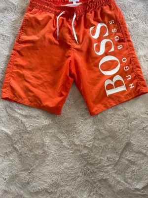  badbyxor från Hugo Boss strl S - Snygga orange badbyxor från Hugo Boss med vit dragsko i midjan och stort vitt BOSS-tryck längs ena benet. Klassisk modell med innerbyxa i mesh och elastisk midja. Perfekta för stranden eller poolen.