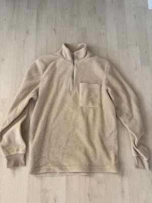 Beige fleece-tröja från Jack & Jones - Mysig beige fleece-tröja från Jack & Jones med halv dragkedja och hög krage. Tröjan har en bröstficka och ribbade muddar vid ärmsluten. Perfekt för lager-på-lager och tillverkad i mjukt teddy-material.
