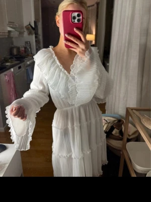 Vit studentklänning från Gina Tricot - Superfin vit studentklänning från Gina Tricot med volangdetaljer och plisserat tyg. Klänningen har långärmade ärmar med volangkant och v-ringad hals. 
