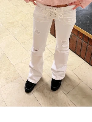 Vita bootcut jeans med snörning - Säljer ett par vita bootcut jeans med snörning i midjan och slitna detaljer på benen. Helt slutsålda där av priset!