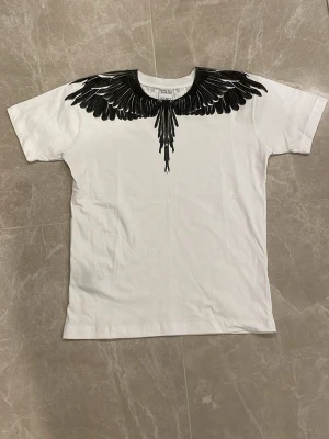 Vit t-shirt med svarta vingar från Marcelo Burlon - Vit t-shirt från Marcelo Burlon med ikoniskt svart vingmönster runt halsen både fram och bak. Klassisk passform och rund halsringning. Perfekt för dig som vill sticka ut med en unik design.
