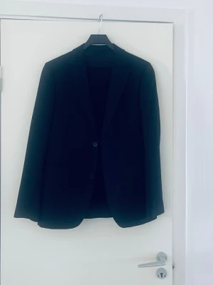 Svart kostym från Riley - Stilren svart kavaj från Riley med klassisk passform och två knappar framtill. Perfekt för en dressad look. Tillverkad i ett mjukt material med slät finish och helfodrad insida.