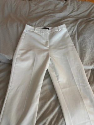 Vita kostymbyxor från Zara - Snygga vita kostymbyxor från Zara med klassisk passform och pressveck. Byxorna har hällor för bälte och stängs med dragkedja, hake och knapp. Perfekta för en stilren look och lätta att matcha med olika toppar.