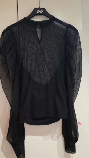 Svart meshblus med puffärmar - Trendig svart blus i mesh med långa puffärmar och rund halsringning. Blusen har transparenta partier och breda muddar vid ärmsluten. Perfekt för dig som vill sticka ut med en edgy och modern look.