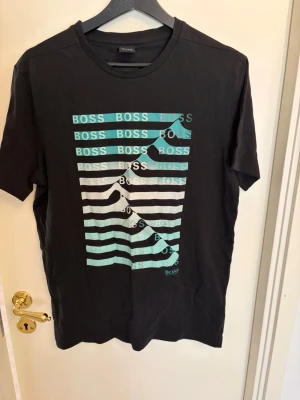 Svart Hugo BOSS t-shirt - Svart t-shirt från BOSS med korta ärmar och rund hals. Framsidan har ett grafiskt tryck med BOSS-loggan i turkos och vitt i ett randigt mönster. Skön passform och tillverkad i mjuk bomull, perfekt för en stilren och modern look.
