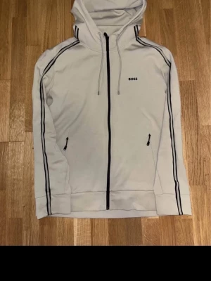 Beige hoodie med svarta detaljer BOSS - Äkta beige tracksuit från BOSS med dragkedja framtill. Utrustad med luva och dragsko samt två fickor med dragkedja och sedan ett bar byxor med. Nästan oanvänd och fräsch. Pris kan diskuteras i DM.