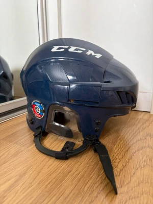Mörkblå hockeyhjälm från CCM - Säljer en mörkblå hockeyhjälm från CCM med visir och justerbart hakskydd. Hjälmen har en rundad form och är tillverkad i hård plast. Märke och logga syns tydligt på sidan och ovansidan. Perfekt för dig som spelar ishockey eller samlar på sportutrustning.