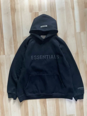Svart Essentials hoodie med tryck - Svart hoodie från Essentials med stor logga på bröstet och mindre logga på huvan. Klassisk känguruficka framtill och ribbade muddar. Oversized passform och mjukt material, perfekt för en avslappnad streetwear-look. Sitter som S