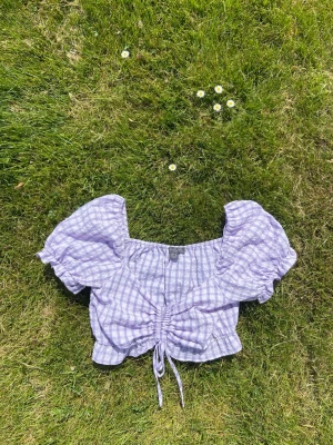 Lila rutig croppad blus med snörning - Supersöt lila och vit rutig blus med puffärmar och croppad passform. Toppen har snörning framtill som ger en snygg rynkeffekt och v-ringad hals. SOOOOMMAAAR BABY 💜🤍💜🤍