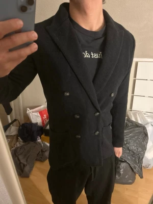 John Henric dubbelknäppt kavaj - Snygg svart kavaj med dubbelknäppning och breda slag. Kavajen har två framfickor och en figurnära passform som ger en stilren look. Perfekt för dig som vill ha en klassisk men modern stil. Materialet ser ut att vara mjukt och bekvämt.