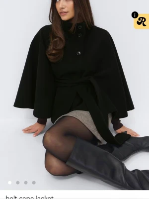 Svart cape-kappa med knytband - Stilren svart cape-kappa med knytband i midjan och stora knappar framtill. Kappan har en lös, svepande passform och hög krage. Perfekt för dig som vill ha en trendig och elegant look. Materialet ser ut att vara ullblandning.