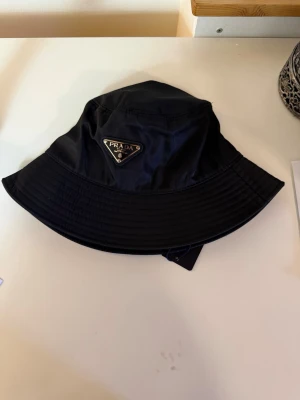 Svart bucket hat från Prada - Stilren svart bucket hat från Prada med klassisk triangellogga framtill. Hatten är tillverkad i glansigt nylonmaterial och har bred brätte. Perfekt accessoar för en trendig streetwear-look.