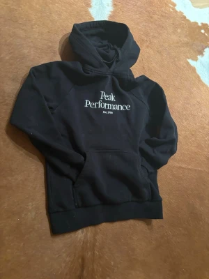Svart hoodie från Peak Performance - Snygg svart hoodie från Peak Performance med vit broderad logga och text 'Est. 1986' på bröstet. Klassisk känguruficka framtill och stor huva. Perfekt för en avslappnad och sportig stil. Mjuk och skön bomullsblandning.