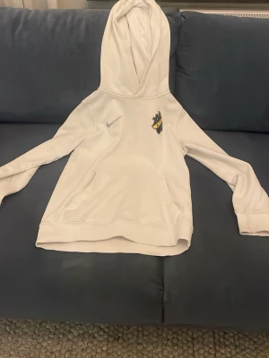 Vit AIK hoodie från Nike - Vit hoodie från Nike med AIK-logga på bröstet och klassisk känguruficka. Mjuk insida och huva för extra komfort. Perfekt för dig som vill visa ditt AIK-stöd med stil. Passar dig som är 137-147 cm lång.