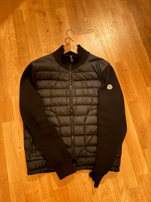 Svart quiltad pufferjacka Moncler - Snygg svart pufferjacka från Moncler med quiltad front och ärmar i mjukare material. Jackan har dragkedja framtill, ribbad krage och Moncler-logga på ärmen. Perfekt för kyliga dagar när du vill ha både stil och komfort.