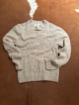Beige stickad tröja från Arket - Snygg beige stickad tröja från Arket med diskreta svarta och grå spräckliga detaljer. Tröjan har rund halsringning, långa ärmar och breda ribbade muddar vid ärmslut och nederkant. Perfekt för lager-på-lager-stil.