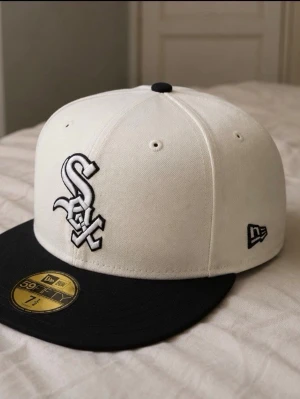Vit och svart White Sox keps New Era - Snygg keps från New Era med White Sox-logga framtill och texten 'White Sox' broderat bak. Kepsen är vit med svart skärm och svart knapp på toppen. Klassisk 59FIFTY-modell med platt skärm, perfekt för dig som gillar sportig streetstyle.