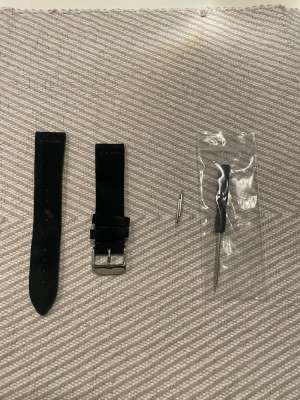 Svart klockarmband i mocka, 20MM - Säljer ett svart klockarmband i mocka med silverfärgat spänne. Paketet innehåller två delar av armbandet, två fästen och ett litet verktyg för enkel montering. Perfekt för att ge din klocka en ny look.