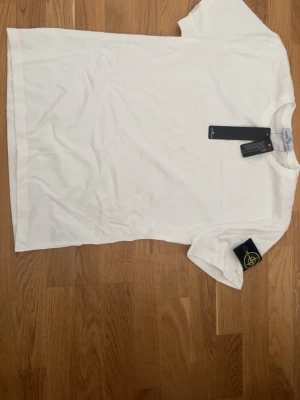 Vit t-shirt från Stone Island - Vit t-shirt från Stone Island med klassisk rund hals och korta ärmar. T-shirten har den ikoniska Stone Island-loggan på vänster ärm och är tillverkad i mjuk bomull. Perfekt basic-plagg med snygg streetwear-känsla.