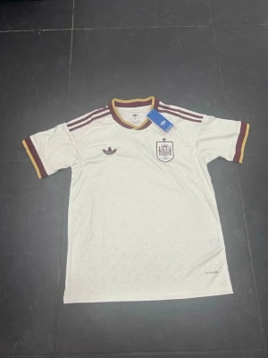 Vit Spanien fotbollströja Adidas - Snygg vit fotbollströja från Adidas med Spaniens landslagsemblem på bröstet och vinröda detaljer. Tröjan har klassiska Adidas-ränder på axlarna, rund halsringning och korta ärmar. Tillverkad i lätt och ventilerande material, perfekt för träning eller match.
