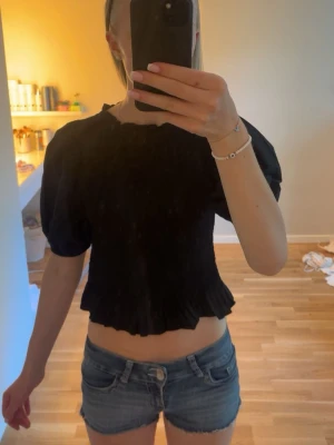 Svart puffärm topp med smock - Trendig svart topp med puffärmar och smockad kropp. Toppen har korta ärmar och en croppad passform med volangkant nedtill. Perfekt för dig som gillar en stilren men ändå lekfull look. Endast provad tvättas innan jag skickar den 