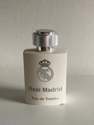 Real Madrid Eau de Toilette parfym - Snygg parfymflaska från Real Madrid i vit färg med silverdetaljer och klubbens logga på framsidan. Flaskan har en robust, fyrkantig form och ett stilrent metallock. Perfekt för dig som vill visa ditt stöd för laget och samtidigt dofta fräscht.