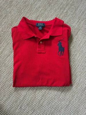Röd pikétröja från Polo Ralph Lauren - Klassisk röd pikétröja från Polo Ralph Lauren med stor mörkblå logga broderad på bröstet. Tröjan har krage, knappar framtill och är gjord i mjuk bomull. Har en mörkblå 3 på höger ärm. Perfekt till golfbanan eller till en stilren look nu till sommaren.