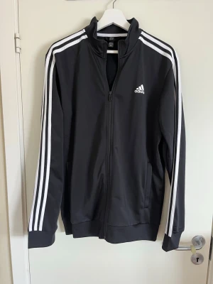 Svart Adidas träningsjacka - Säljer en svart träningsjacka från Adidas med klassiska vita ränder längs ärmarna och vit Adidas-logga på bröstet. Jackan har dragkedja framtill, hög krage och muddar vid ärmslut och nederkant. Perfekt för gymmet eller till vardags.
