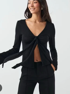 Svart långärmad topp med knyt - Trendig svart långärmad topp från Gina Tricot med djup v-ringning och knyt framtill. Modellen är croppad och har en figurnära passform som framhäver midjan. Perfekt för dig som vill ha en stilren men ändå unik look.