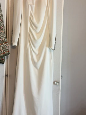 Vit långklänning med strassdetaljer - Elegant vit långklänning med långa ärmar och draperad framsida. Ärmsluten är dekorerade med stora, gnistrande strass och klädda knappar för en lyxig känsla. Perfekt för speciella tillfällen där du vill sticka ut. Pris går att diskutera. Vill bli av med klänningar därför