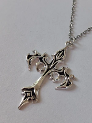 Silvrigt halsband med korshänge - Snyggt halsband med ett stort, detaljerat korshänge i silverfärgad metall. Hänget har en unik, ornamenterad design på båda sidorna av den, med svarta inslag som ger en cool och edgy look. Kedjan är tunn och matchar hängets färg. Materialet rostfritt stål 