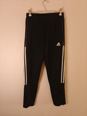 Adidas träningsbyxor - Säljer ett par svarta träningsbyxor från Adidas med vita ränder längs sidorna och Adidas-logga på låret. Byxorna är tillverkade i Primegreen-material och har en smal passform med resår i midjan. Perfekta för träning eller chill.