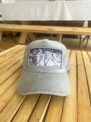Ljusgrå keps från Berrettio Sweden med Cristiano Ronaldo - Säljer en ljusgrå keps från Berrettio Sweden med mesh baktill och justerbar rem. Framtill finns ett svartvitt tryck med motiv av en person och en pokal. Kepsen har böjd skärm och klassisk trucker-modell. Perfekt för dig som vill sticka ut med en unik accessoar.
