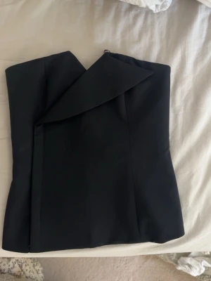 Svart axelbandslös tubtopp - Snygg svart axelbandslös tubtopp med unik omlottdetalj framtill. Toppen har en stilren och modern look som passar perfekt till jeans eller kjol. Materialet känns mjukt och följsamt mot kroppen och ger en elegant siluett.