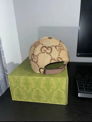Beige GG keps från Gucci ÄKTA - Säljer en beige keps från Gucci med det klassiska GG-mönstret i brunt. Kepsen har justerbar rem bak och är tillverkad i canvasmaterial. Perfekt för dig som vill ha en stilren och lyxig accessoar med tydlig logga.