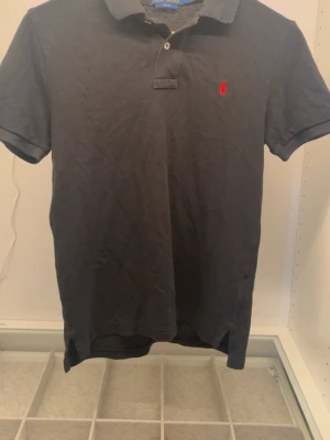 Svart pikétröja från Ralph Lauren - Klassisk svart pikétröja från Ralph Lauren med korta ärmar och den ikoniska röda logotypen broderad på bröstet. Tröjan har krage, knappar vid halsen och är tillverkad i mjuk bomull. Perfekt till både jeans och shorts.