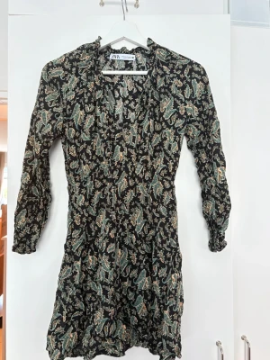 Svart mönstrad klänning från Zara - Svart klänning från Zara med grönt och orange paisleymönster. Klänningen har långa ärmar, knytning vid halsen och lätt rynkade detaljer. 
