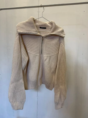 Beige stickad tröja från Gina Tricot - Säljer en beige stickad tröja från Gina Tricot med halv dragkedja framtill och bred krage. Tröjan har grovstickad struktur, långa ärmar och ribbade muddar. Perfekt för dig som gillar en avslappnad och trendig look.