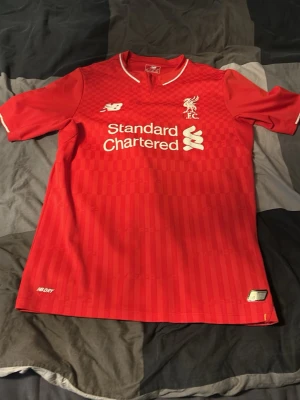 Liverpool FC matchtröja röd New Balance - Säljer en röd Liverpool FC matchtröja från New Balance med vita detaljer och klubbmärke på bröstet. Tröjan har tryckt sponsorlogga och klassisk krage med vit kant. Tillverkad i lätt och ventilerande material, perfekt för fotbollsträning eller match.