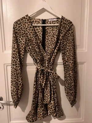 Leopardmönstrad omlottklänning NA-KD - Snygg kort leopardmönstrad omlottklänning från NA-KD med långa ärmar och knyt i midjan. Klänningen har v-ringning och är perfekt för dig som vill sticka ut med ett djurmönster. Tillverkad i ett mjukt och lätt material.