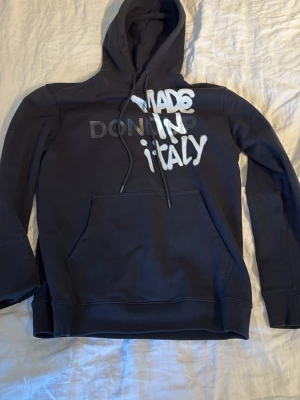Svart hoodie med tryck 'MADE IN ITALY' - Svart hoodie med stor ficka framtill och huva med dragsko. På bröstet står det 'MADE IN ITALY' i vit och grå text med street-inspirerad stil. Mjuk bomullskänsla och ribbade muddar vid ärmar och nederkant. Perfekt för en avslappnad look.