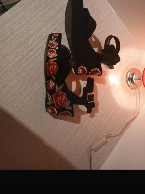 4 par jättefina klackskor - Unika svarta sandaler med platå och broderade röda rosor på sidorna. Sandalerna har en öppen tå, breda remmar och justerbart spänne i guld. Materialet ser ut att vara mocka eller liknande tyg. Perfekta för dig som vill sticka ut med snygga detaljer.