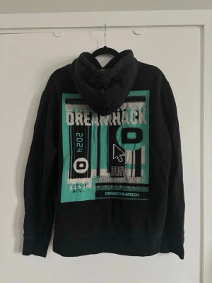 Svart DreamHack hoodie med tryck - Svart hoodie från DreamHack 2024 med stort grafiskt tryck i turkos, vitt och svart på ryggen och mindre logga på bröstet. Klassisk huva med dragsko och magficka framtill. 