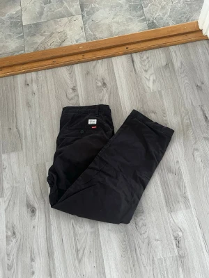 Svarta chinos från Levi's - Säljer ett par svarta chinos från Levi's med klassisk rak passform. Byxorna har knappgylf, sidofickor och bakfickor med knapp. Tillverkade i ett mjukt och slitstarkt bomullstyg som är sköna att bära hela dagen.