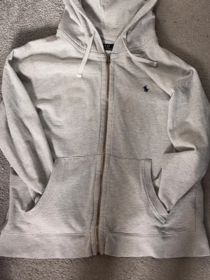 Grå zip hoodie från Polo Ralph Lauren - Snygg ljusgrå hoodie med dragkedja från Polo Ralph Lauren. Klassisk design med huva, dragsnören och två stora fickor framtill. Broderad mörkblå logga på bröstet. Tillverkad i mjukt bomullsmaterial för skön känsla.