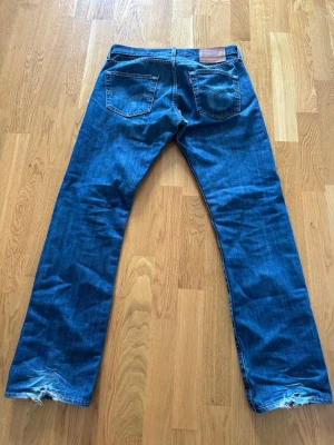 Blå raka Levi's jeans - Säljer ett par klassiska blå Levi's jeans med raka ben och femficksmodell. Jeansen har slitna detaljer vid bensluten och är tillverkade i robust denim. Perfekta för en avslappnad och tidlös stil.